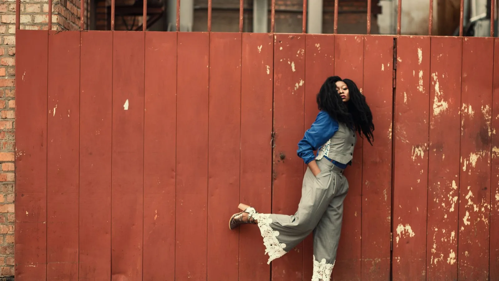 Fashionable woman posing against a vibrant red gate in an urban setting, showcasing modern style.